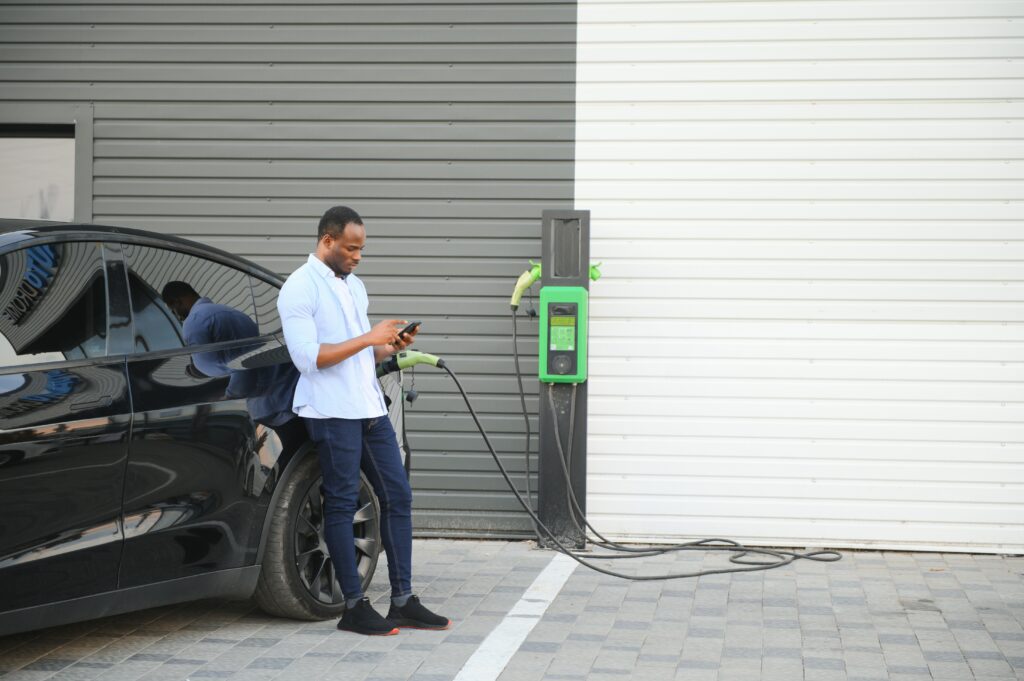 An African man is standing near an electric car, waiting for it to charge at a charging station and using the phone Boca Electrical Services Boca Raton