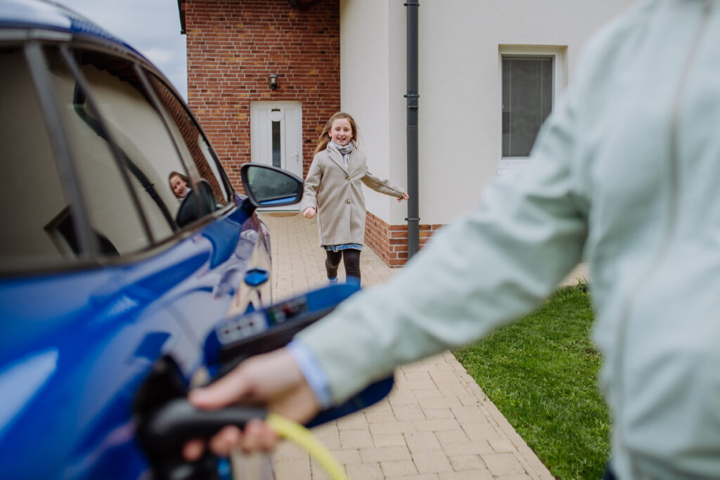 Close up of man holding power supply cable and charging his new electric car, his daughter running to him. Boca Electrical Services Boca Raton