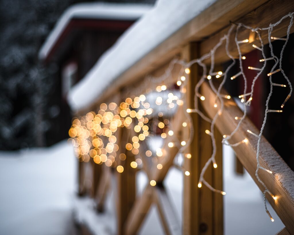 A closeup of Christmas lights on a wooden building covered in the snow on a blurry background