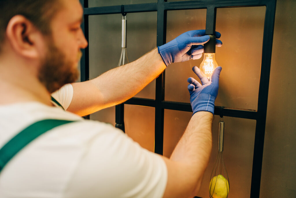 Electrician in uniform replaces the light bulb, handyman. Professional worker makes repairs around the house, home repairing service