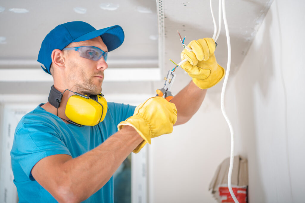 Closeup of Electrician in Protective Gloves and Glasses Doing Illumination Installation Work Stripping Cables with Professional Wire Stripper. Residential Building Renovation Work Theme.