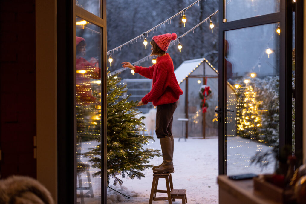 Young woman in red decorates lush Christmas tree with festive ballls and garland at backyard of her house on snow fall, preparing for a winter holidays