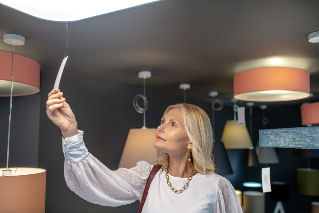 Price tag of a pendant lamp. Successful woman with jewelry on her neck looking up at the price tag of a chandelier, raising her hand, interested.